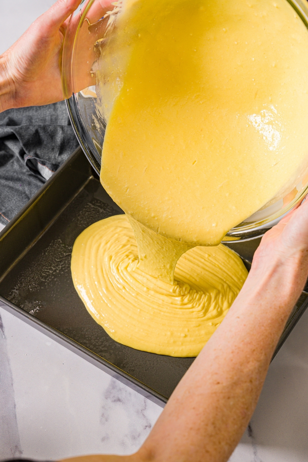 A bowl of yellow cake batter being poured into a greased baking pan. The pan is on a marble counter.