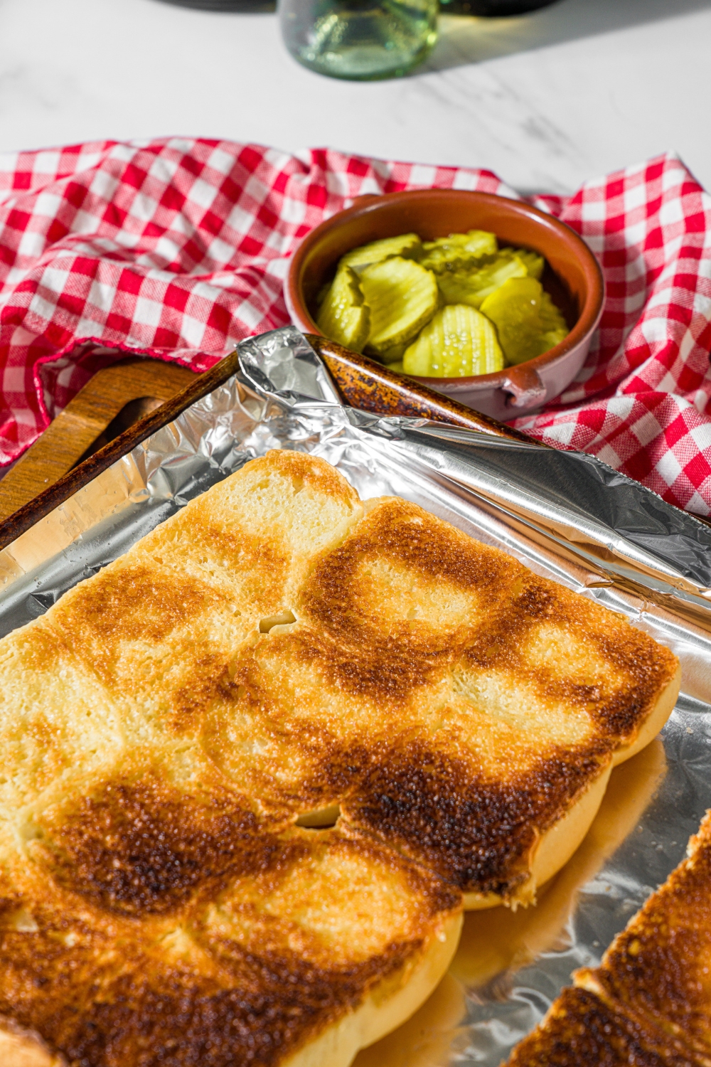 A baking sheet lined with foil with hawaiian rolls sliced in half and toasted. The sheet is on a wooden board with a red checkered napkin and small bowl of pickles.