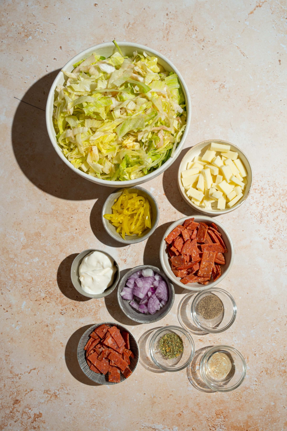 A tan counter with several bowls containing ingredients to make Italian grinder dip including chopped lettuce, pepperoni, salami, provolone cheese, banana peppers, red onion, mayo, and more.