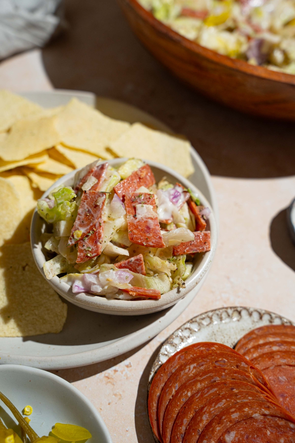 A plate of chips with a small bowl of Italian grinder dip including chopped salami, pepperoni, cheese, banana peppers, and more. The plate is on a tan counter with plates of ingredients and bowl of Italian grinder dip.