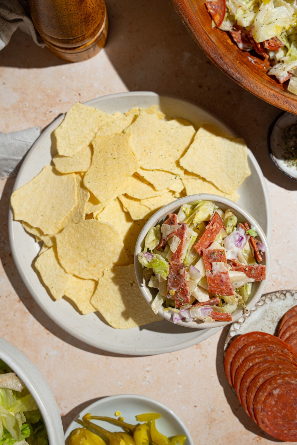 A plate of chips with a small bowl of Italian grinder dip including chopped salami, pepperoni, cheese, banana peppers, and more. The plate is on a tan counter with plates of ingredients and bowl of Italian grinder dip.