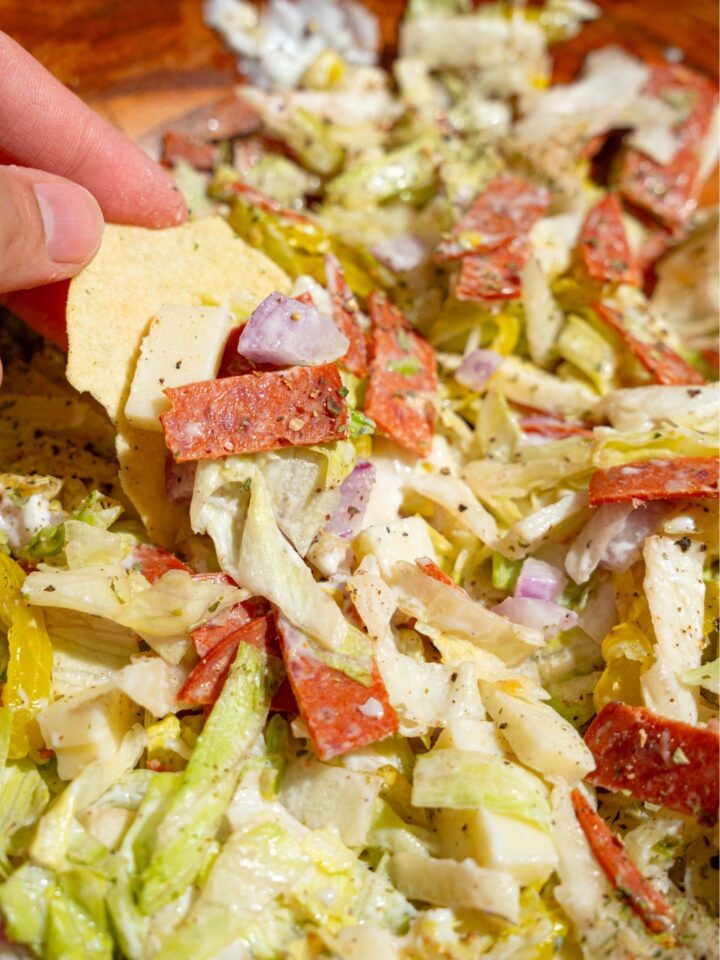 A hand holding a kettle chip scooping up some Italian hoagie dip from a wooden bowl that is filled with the dip.