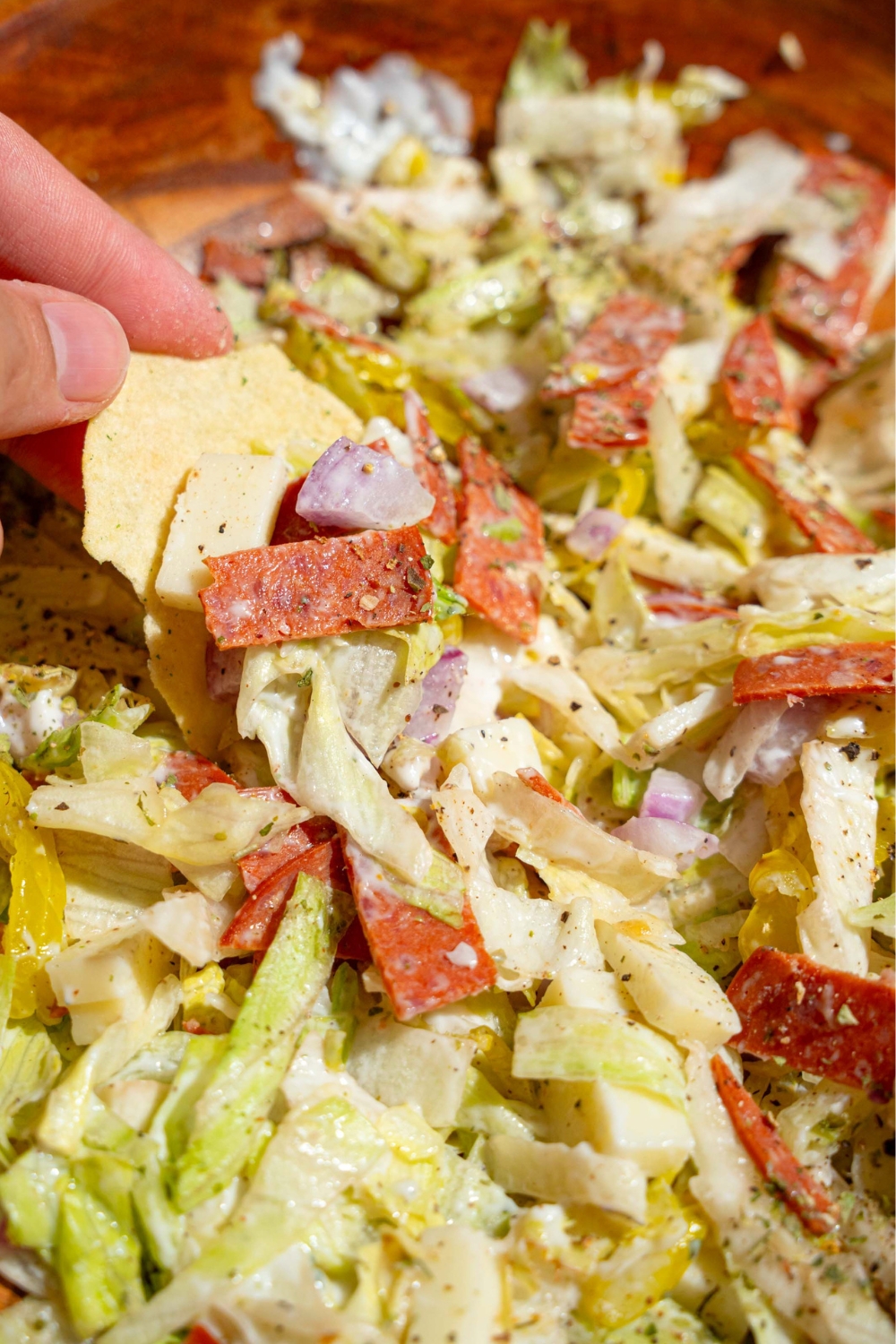 A hand holding a kettle chip scooping up some Italian hoagie dip from a wooden bowl that is filled with the dip.