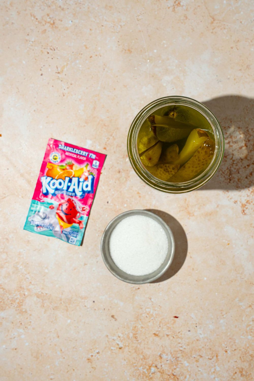 A tan counter with ingredients to make kool aid pickles including a jar of pickles, a packet of kool aid, and bowl of sugar.