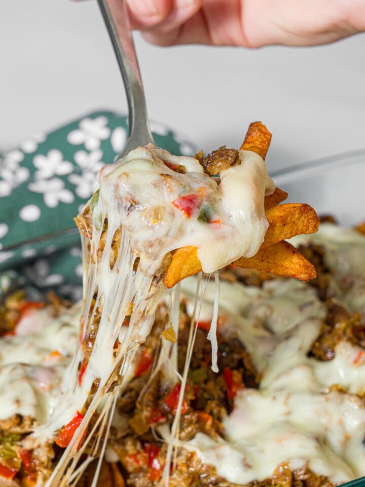 A baking dish with baked Philly cheesesteak loaded fries including fries, melted cheese, and a philly cheesesteak mixture. A fork is pulling a bite of fries from the dish. The dish is on a marble counter with a green floral napkin.