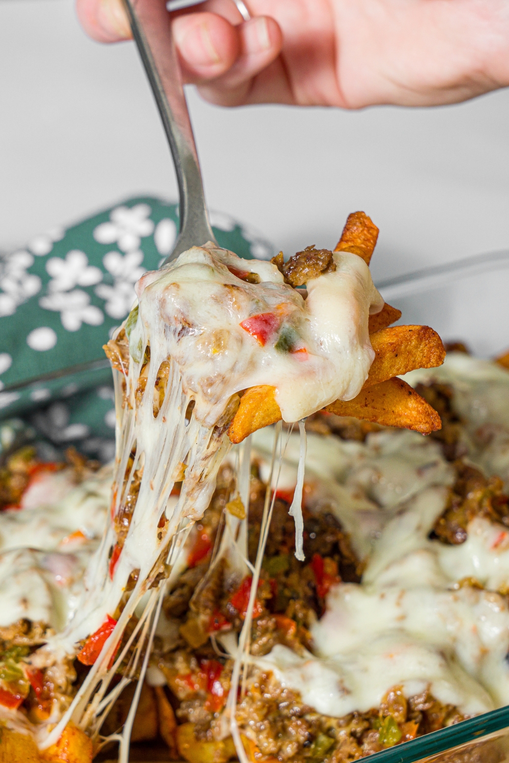 A baking dish with baked Philly cheesesteak loaded fries including fries, melted cheese, and a philly cheesesteak mixture. A fork is pulling a bite of fries from the dish. The dish is on a marble counter with a green floral napkin.