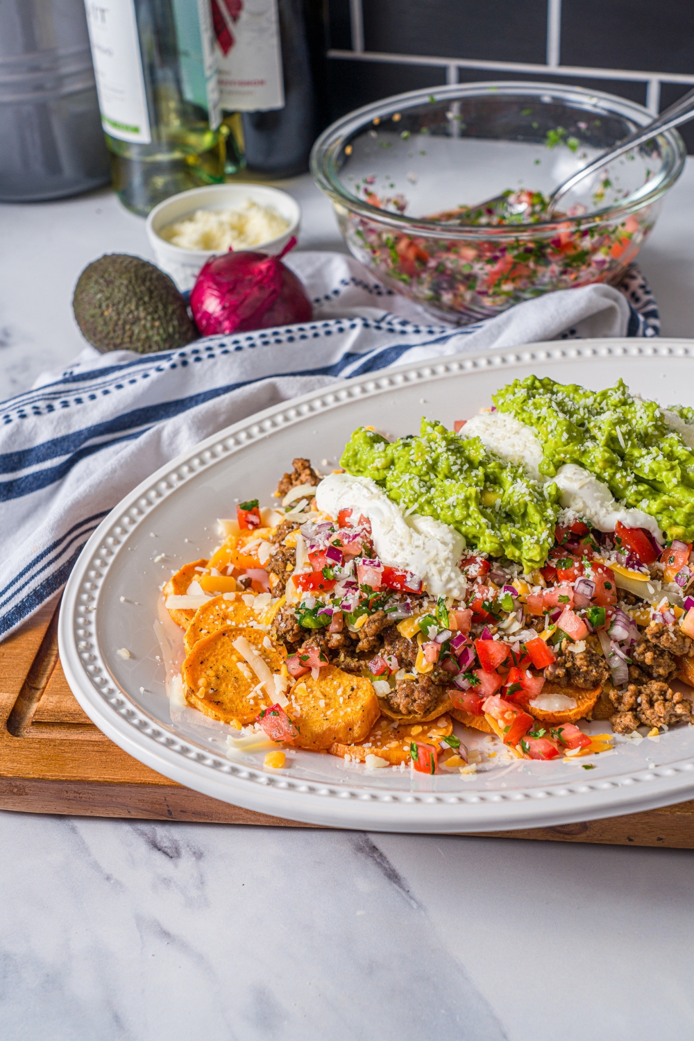A white platter with sweet potato nachos including sweet potato medallions topped with seasoned ground beef, pico de gallo, cheese, sour cream, and guacamole. The platter is on a wooden board on a marble counter with a blue striped napkin.