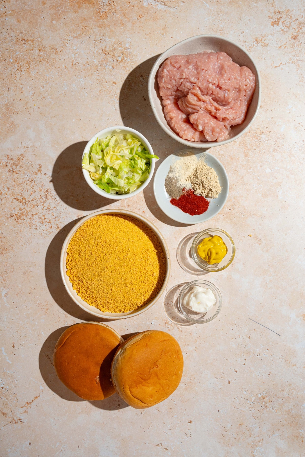 A tan counter with several bowls containing ingredients to make a McChicken sandwich including ground chicken, crushed cornflakes, shredded lettuce, mayo, mustard, hamburger buns, and seasonings.