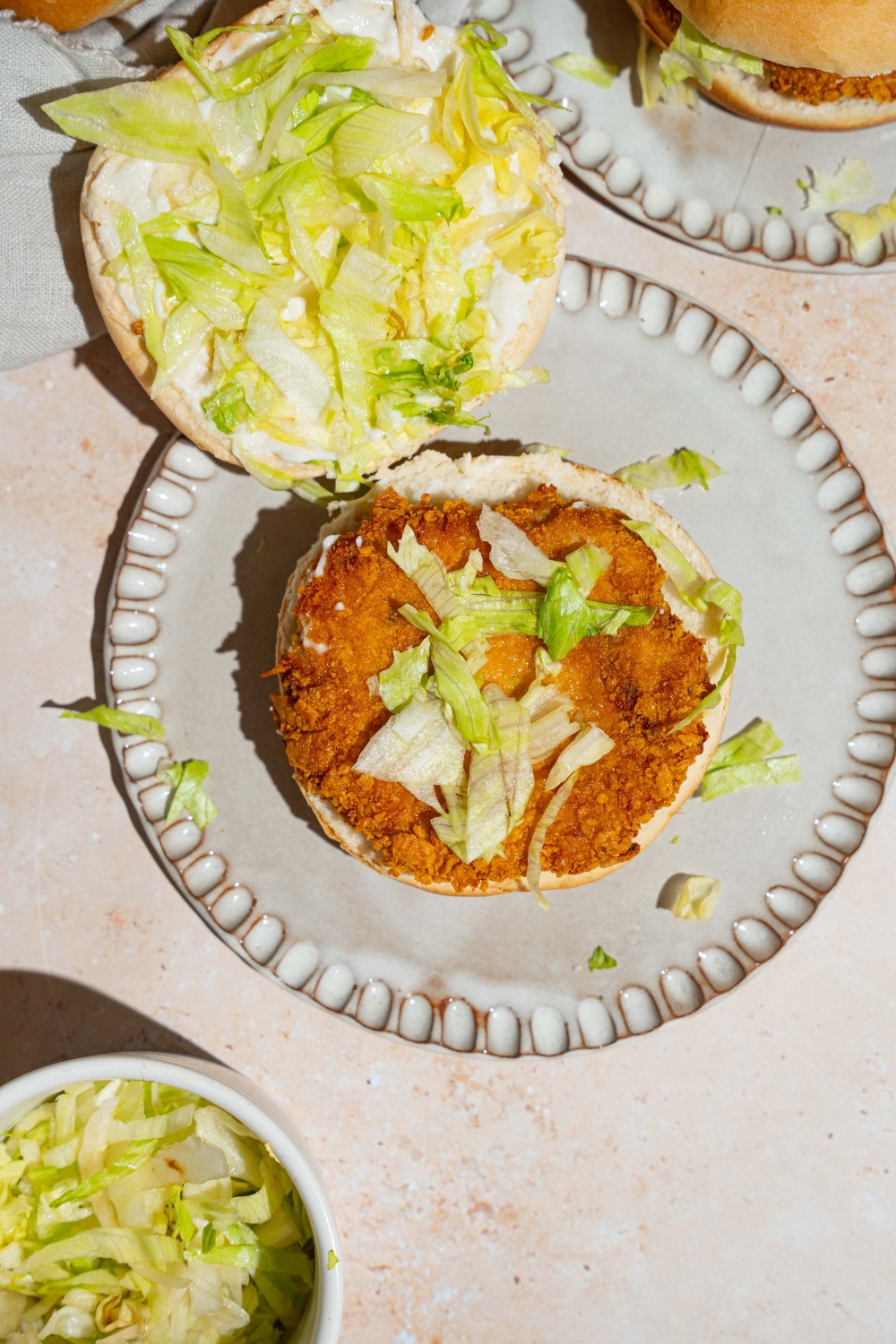 A plate with an open McChicken sandwich topped with shredded lettuce. The plate is on a tan counter with a bowl of shredded lettuce.