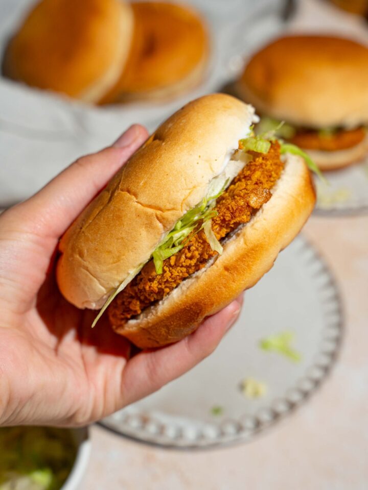 A close up of a hand holding a McChicken sandwich. There is a tan counter with an additional sandwich blurred in the background.