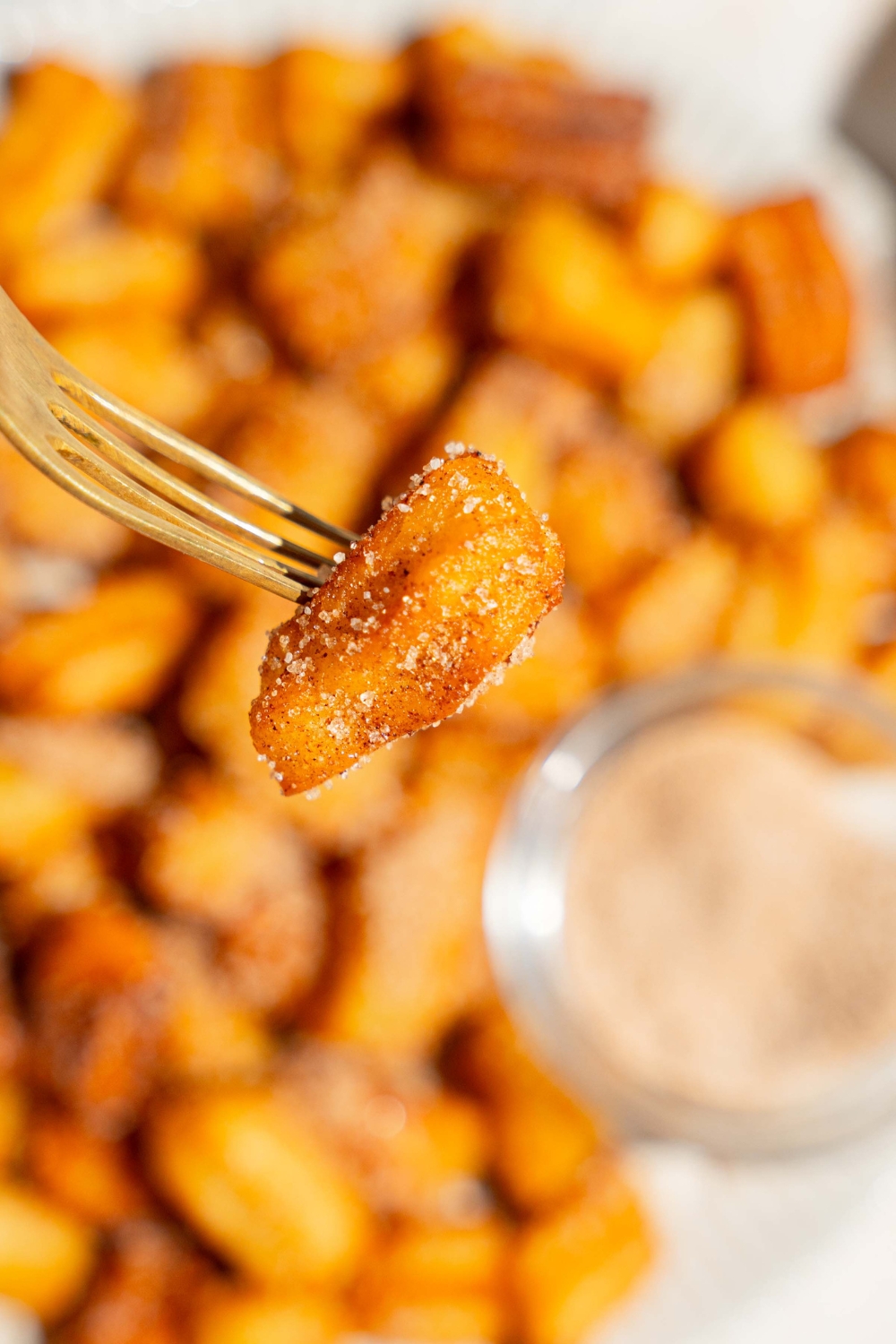 A close up of a fork with a mini churro coated in cinnamon sugar. There is a white plate of mini churros blurred in the background.