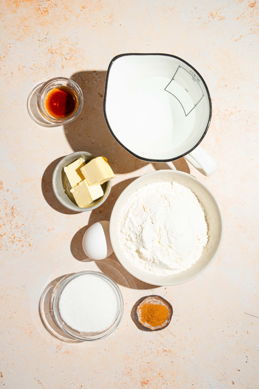 A tan counter with several bowls containing ingredients to make mini churros including flour, butter, vanilla, water, egg, cinnamon, and sugar.