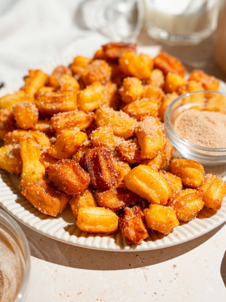 A white plate with mini churros coated in cinnamon sugar. There is a bowl of cinnamon sugar on the plate. The plate is on a tan counter.