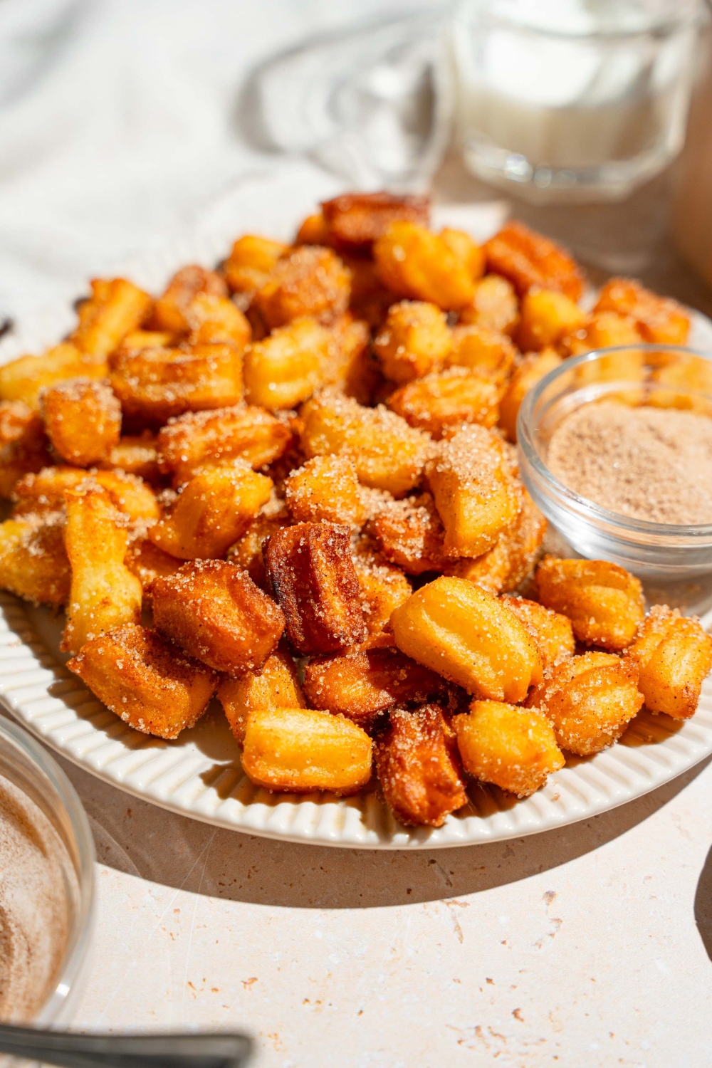 A white plate with mini churros coated in cinnamon sugar. There is a bowl of cinnamon sugar on the plate. The plate is on a tan counter.