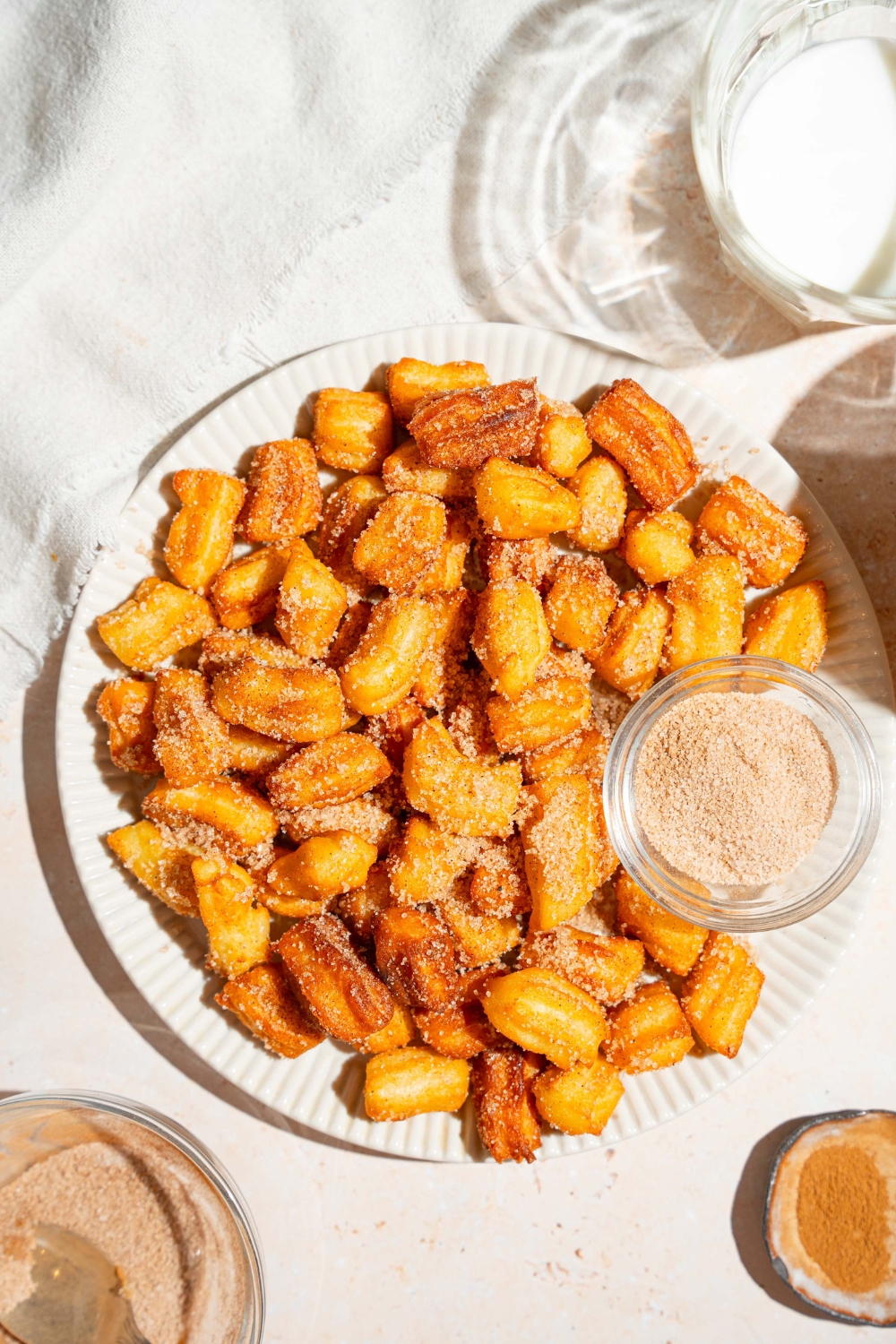 A white plate with mini churros coated in cinnamon sugar. There is a bowl of cinnamon sugar on the plate. The plate is on a tan counter.