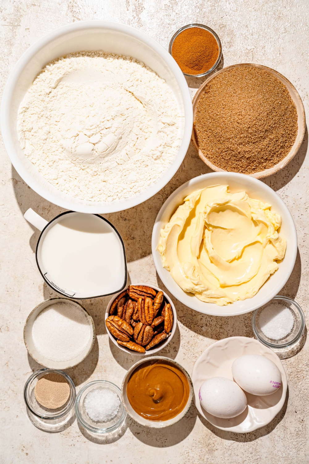 A tan counter with several bowls containing ingredients to make pecan caramel sticky buns including flour, cinnamon, sugar, butter, eggs, brown sugar, caramel sauce, pecans, and sea salt.