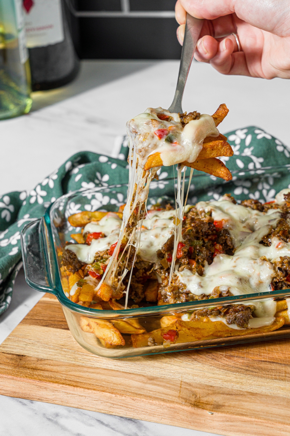A baking dish with baked Philly cheesesteak loaded fries including fries, melted cheese, and a philly cheesesteak mixture. A fork is pulling a bite of fries from the dish. The dish is on a wooden board on a marble counter with a green floral napkin.