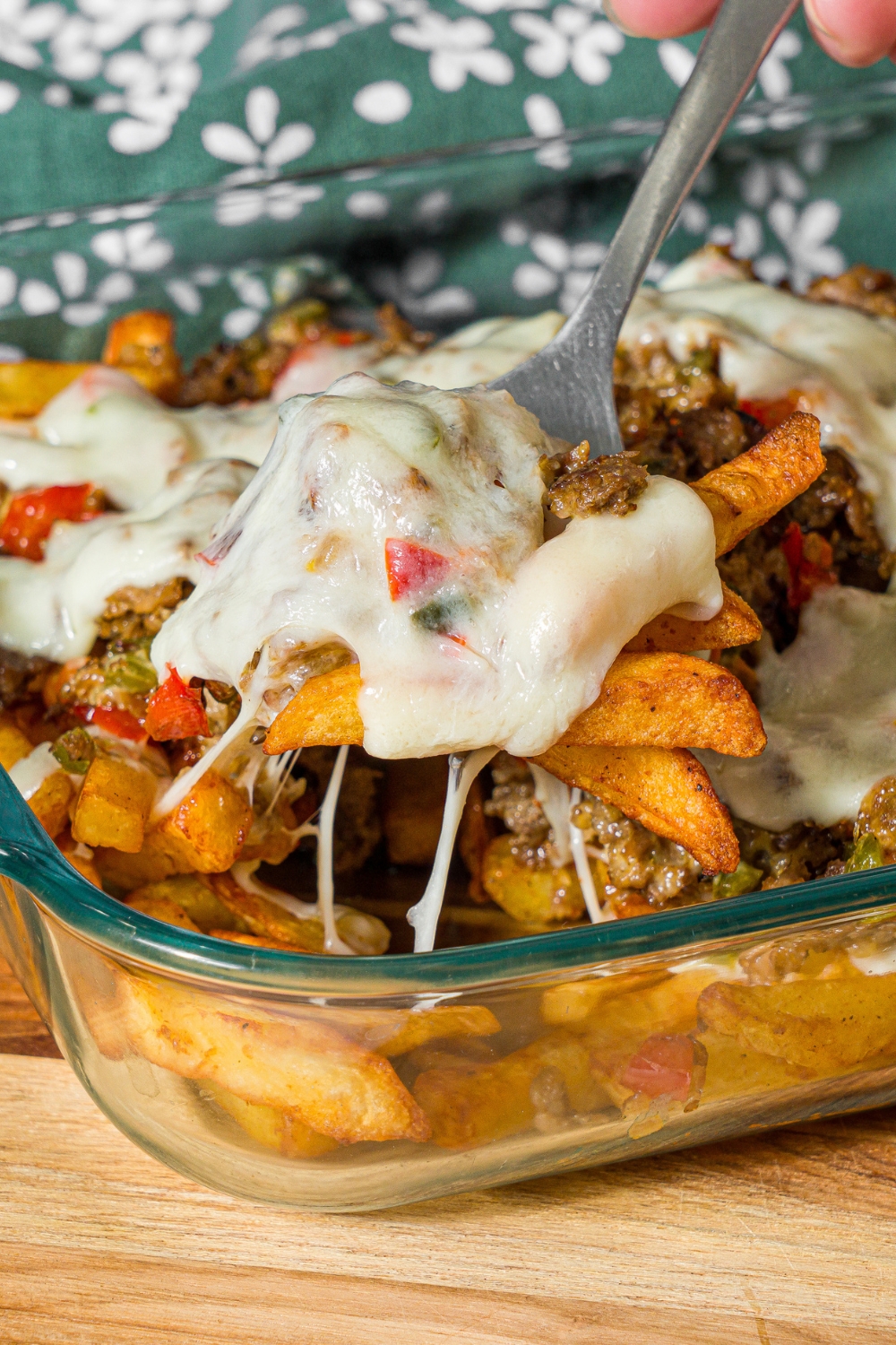 A baking dish with baked Philly cheesesteak loaded fries including fries, melted cheese, and a philly cheesesteak mixture. A fork is pulling a bite of fries from the dish. The dish is on a wooden board with a green floral napkin.