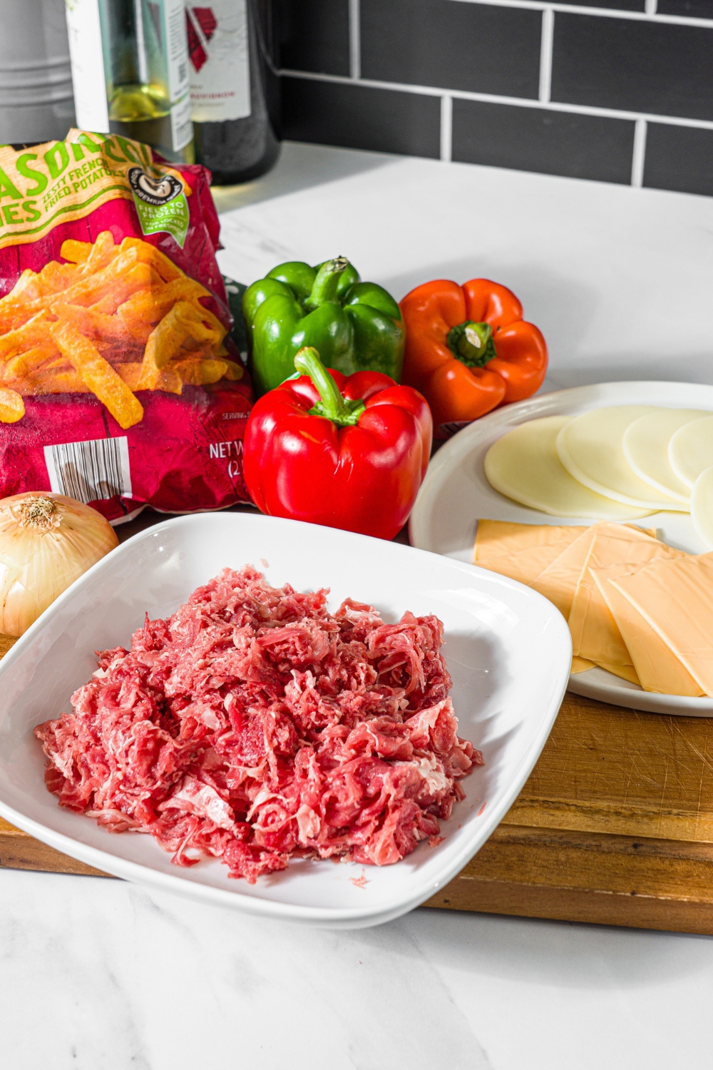A wooden board with ingredients to make philly cheesesteak loaded fries including a bag of fries, ground beef, sliced cheeses, and peppers.