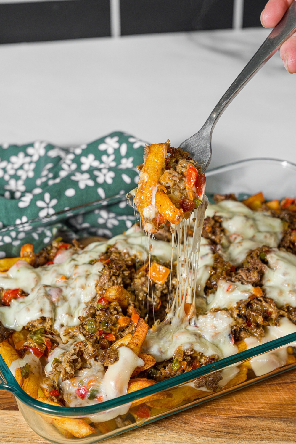 A baking dish with baked Philly cheesesteak loaded fries including fries, melted cheese, and a philly cheesesteak mixture. A fork is pulling a bite of fries from the dish. The dish is on a wooden board.