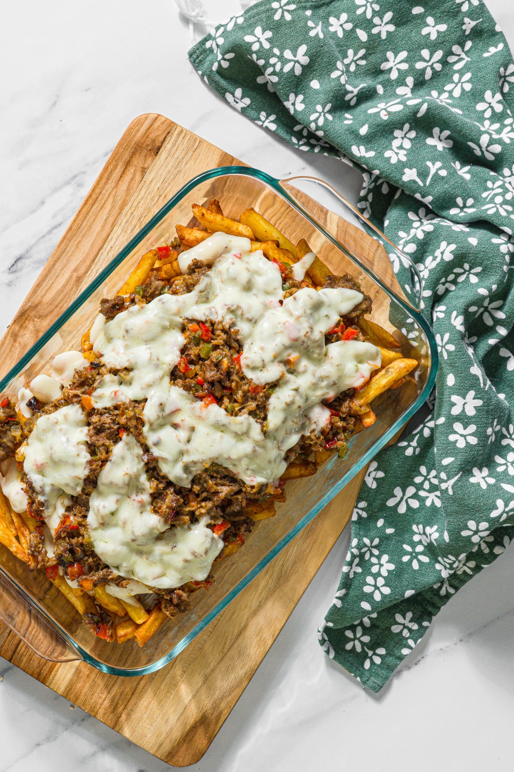 A baking dish with baked Philly cheesesteak loaded fries including fries, melted cheese, and a philly cheesesteak mixture. The dish is on a wooden board on a marble counter with a green floral napkin.