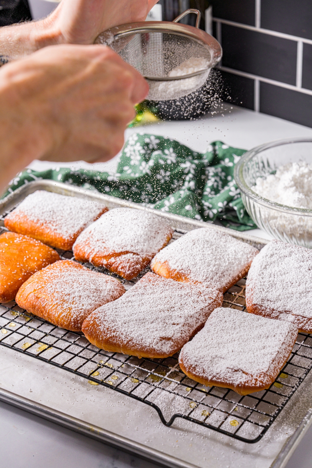 A wire rack with fried beignets with powdered sugar being dusted over them through a sieve. The rack is on a baking sheet lined with parchment paper.