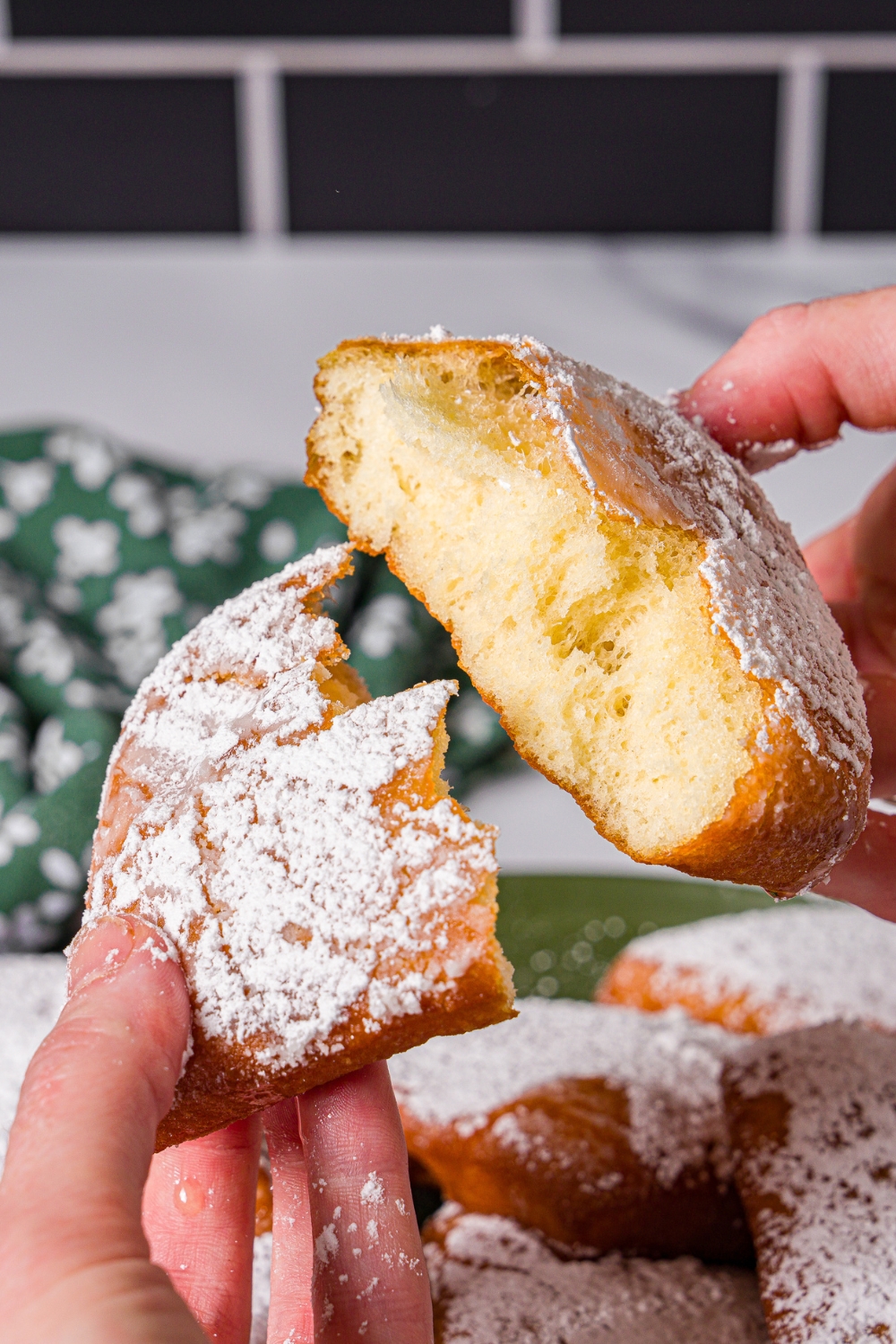 A hand splitting apart a beignet topped with honey and powdered sugar. There is a plate of beignets in the background.