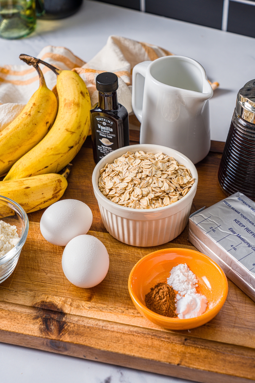 A wooden board with ingredients to make protein banana muffins including bananas, rolled oats, eggs, vanilla, cream cheese, protein powder, syrup, and more.