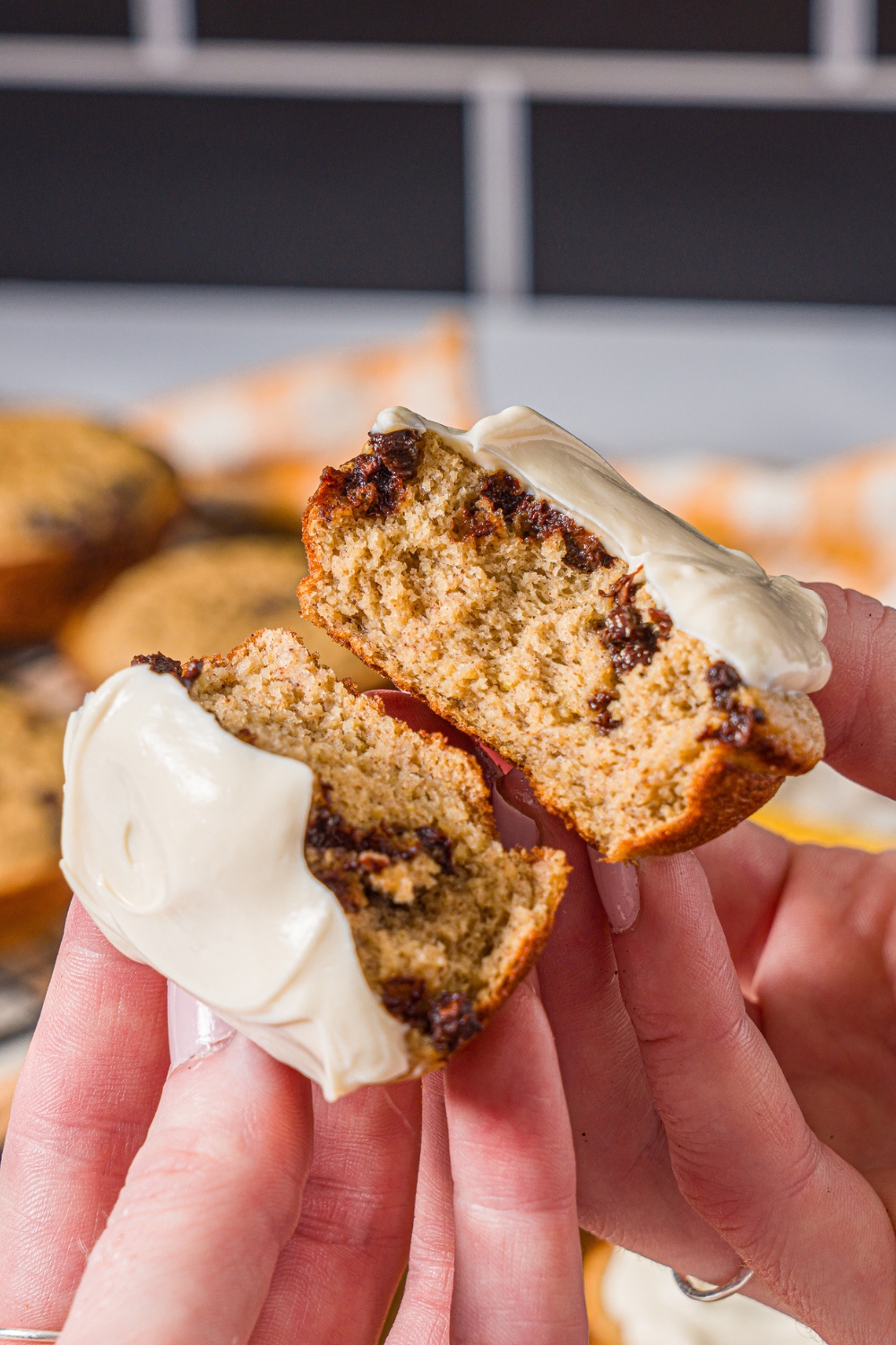 A close up of a hand splitting a frosted protein banana muffin in half. There is a wooden board with a rack of muffins blurred in the background.