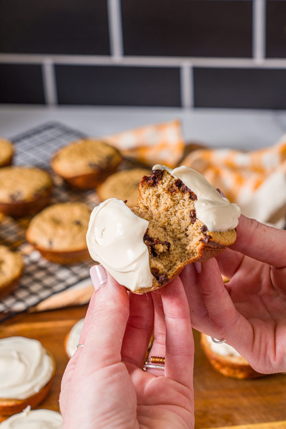 A close up of a hand splitting a frosted protein banana muffin in half. There is a wooden board with a rack of muffins blurred in the background.