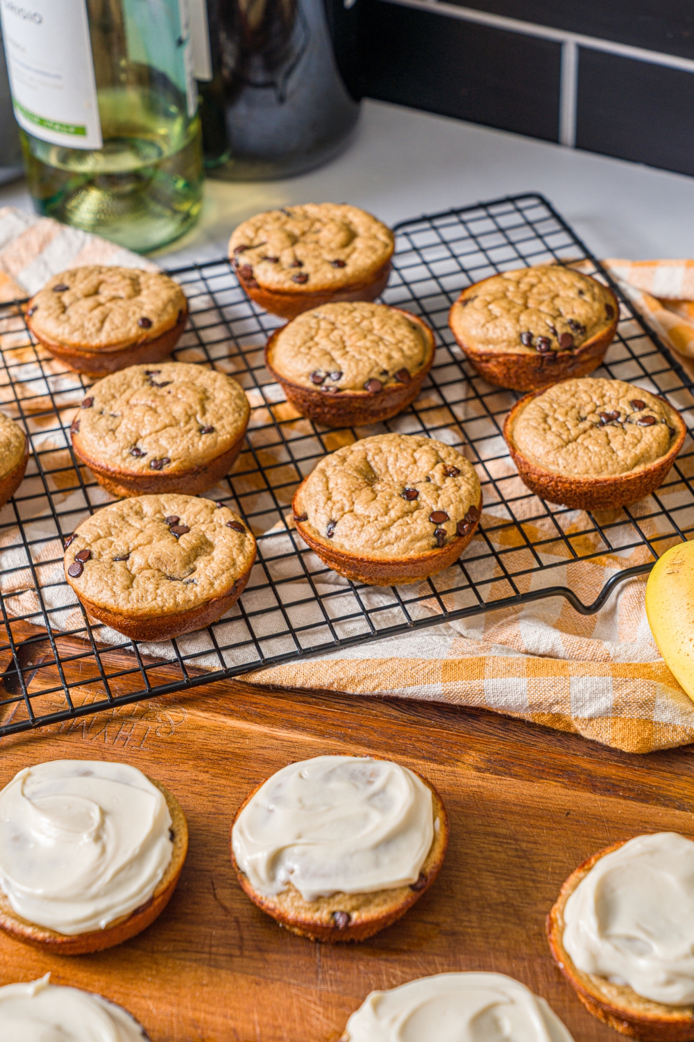 A wire rack with protein banana muffins with a wooden board with frosted muffins. The rack is on a yellow striped napkin.