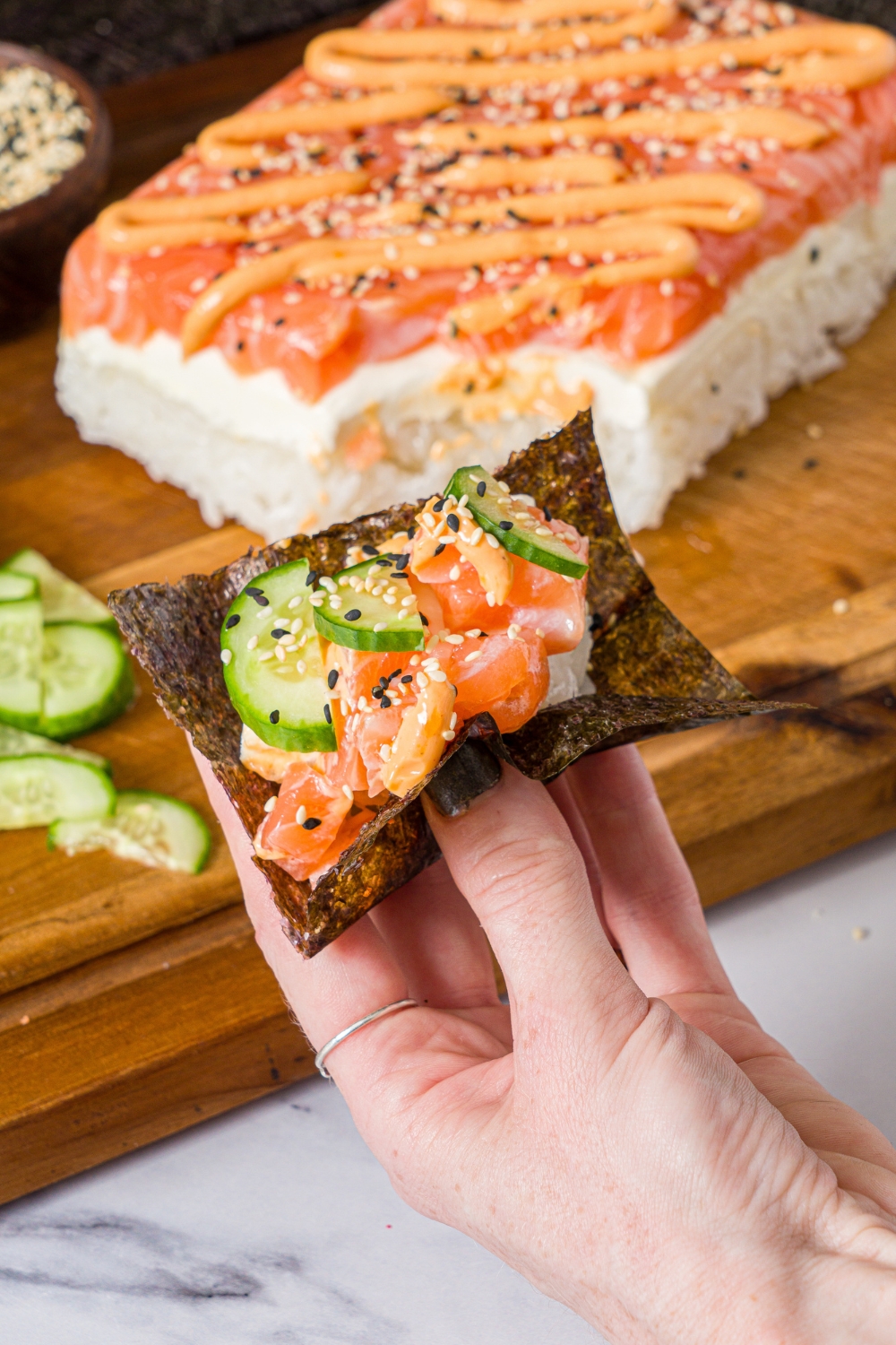 A hand holding a nori sheet stuffed with salmon sushi cake topped with sliced cucumber and sesame seeds. There is a tray of sushi cake on a wooden board in the background.