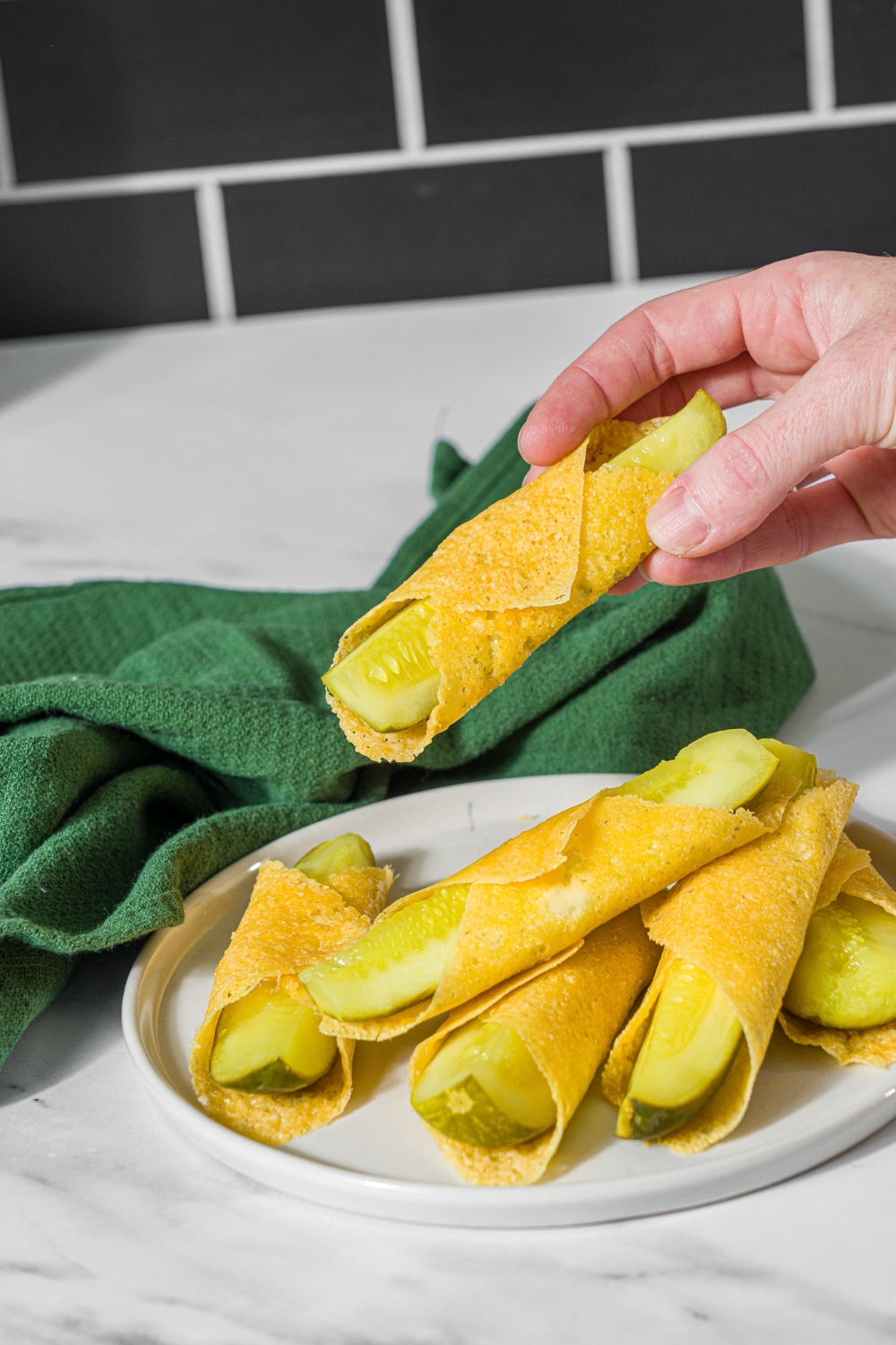 A plate with a pile of chickles made with pickles and cheese. A hand is taking one of the chickles. The plate is on a marble counter with a green cloth napkin.