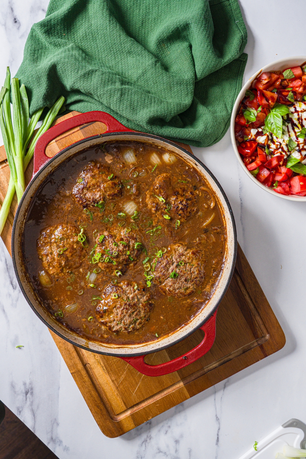 A cast iron skillet with hamburger patties in onion gravy garnished with sliced green onion. The skillet is on a wooden board with a green cloth napkin.