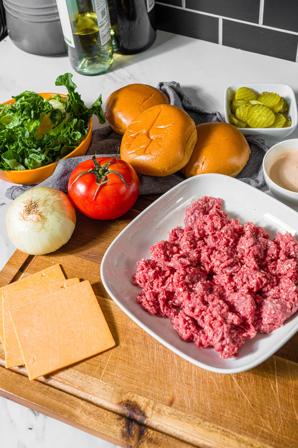 A wooden board with ingredients to make double smash burger including ground beef, burger buns, sliced cheese, onion, tomato, lettuce, pickles, and burger sauce.
