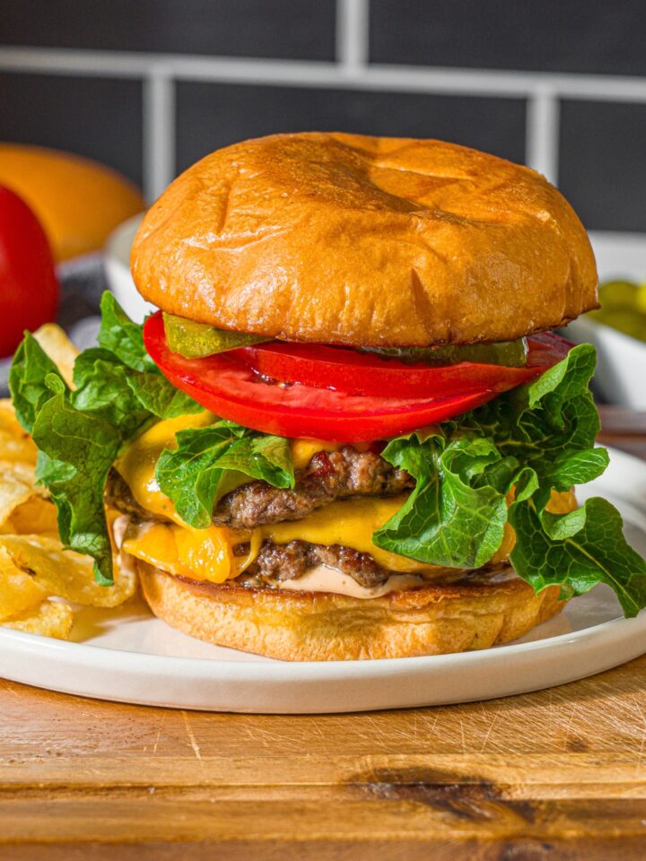 A white plate with a double smash burger with lettuce, caramelized onions, tomato, pickles, and burger sauce served with chips. The plate is on a wooden board.
