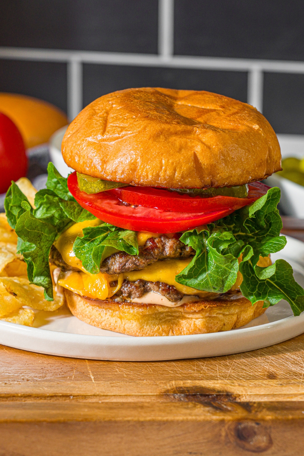 A white plate with a double smash burger with lettuce, caramelized onions, tomato, pickles, and burger sauce served with chips. The plate is on a wooden board.