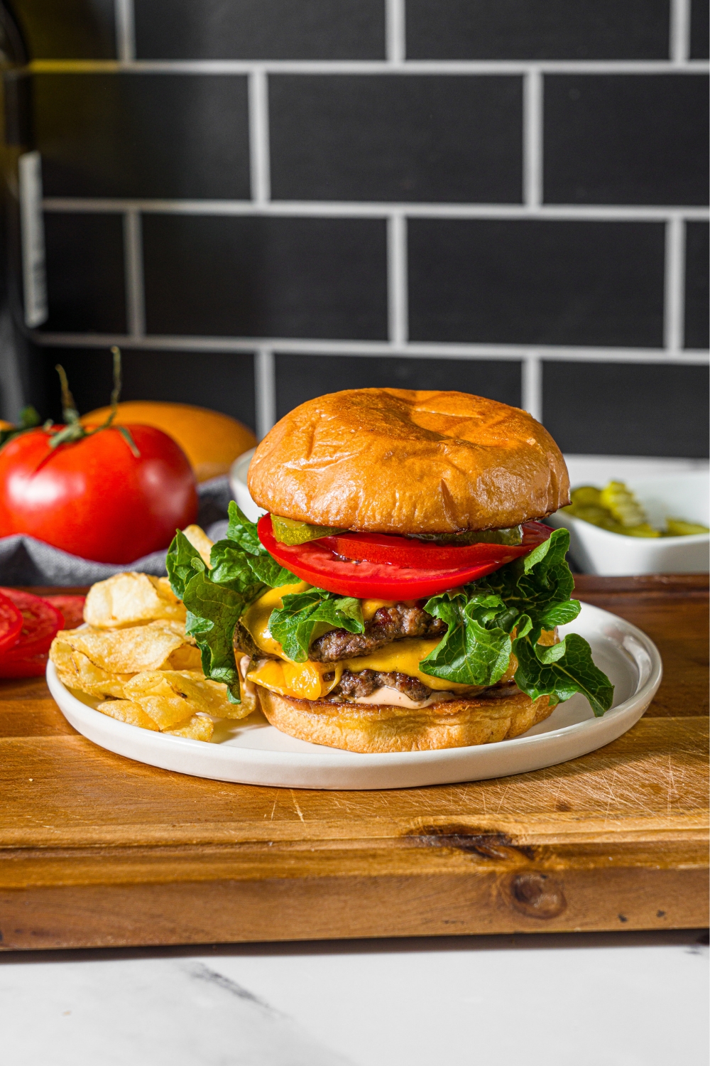 A white plate with a double smash burger with lettuce, caramelized onions, tomato, pickles, and burger sauce served with chips. The plate is on a wooden board.