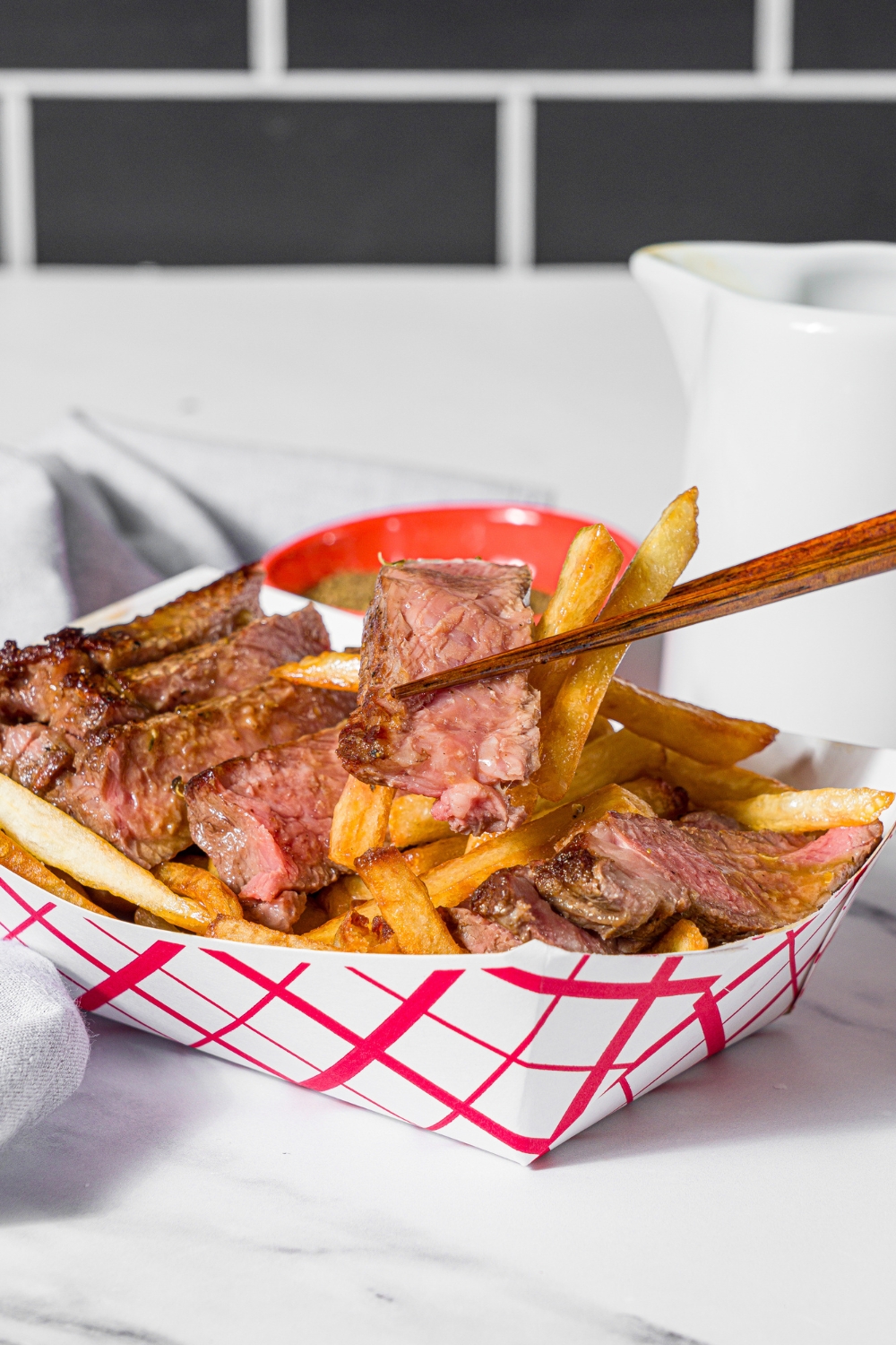 A fry basket with french fries topped with sliced steak and topped with steak frites sauce. A pair of chopsticks are taking a bite of fries. The basket is on a marble counter with a bowl of seasonings and a gray cloth napkin.