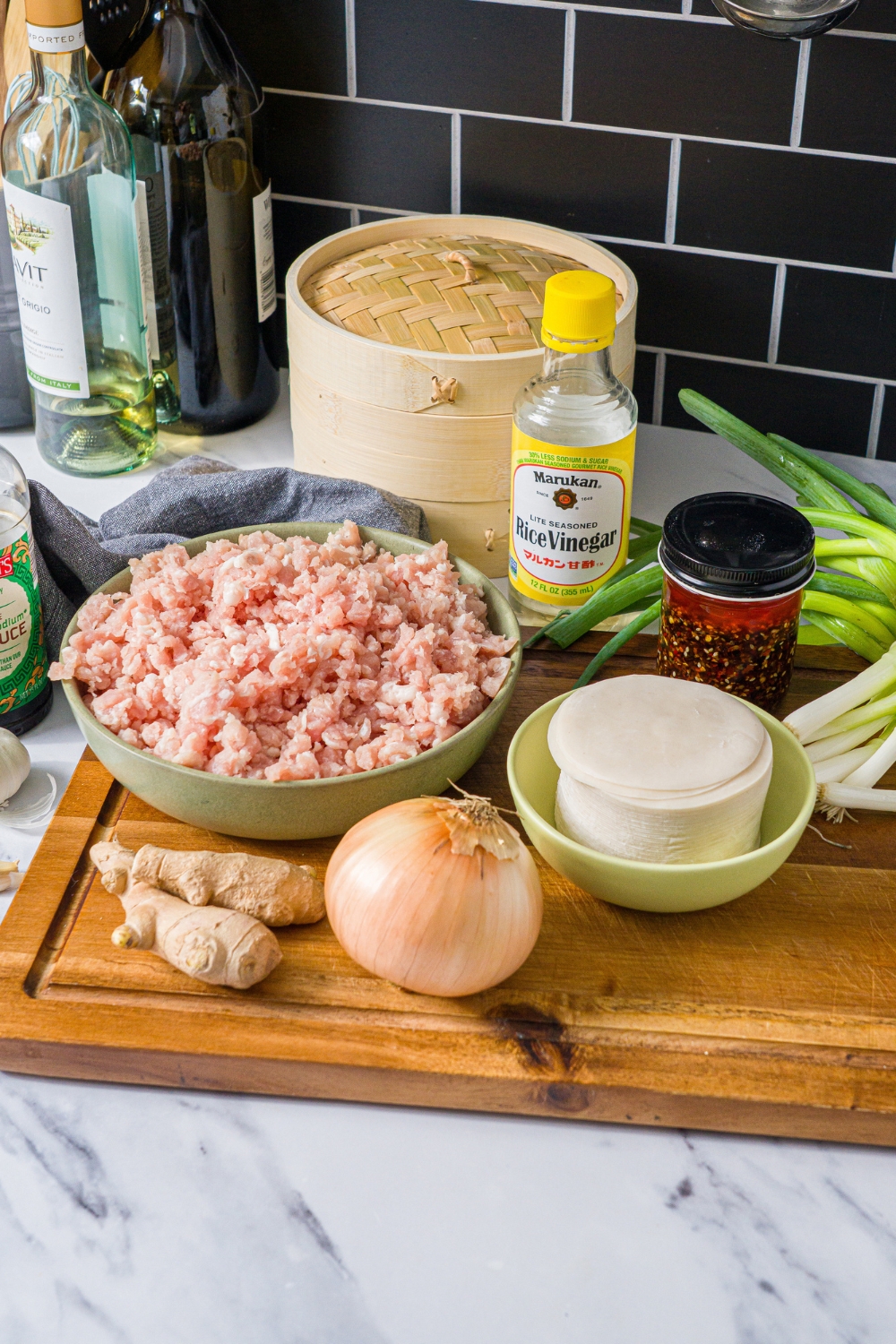 A wooden board with ingredients to make steamed dumplings including ground pork, onion, ginger, dumpling wrappers, rice vinegar, chili crisp, and green onion.