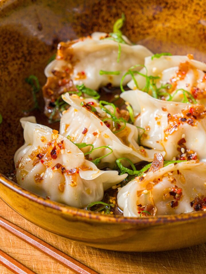 A bowl of steamed pork dumplings in chili oil topped with sliced green onion. The bowl is on a wooden board with a pair of chopsticks.