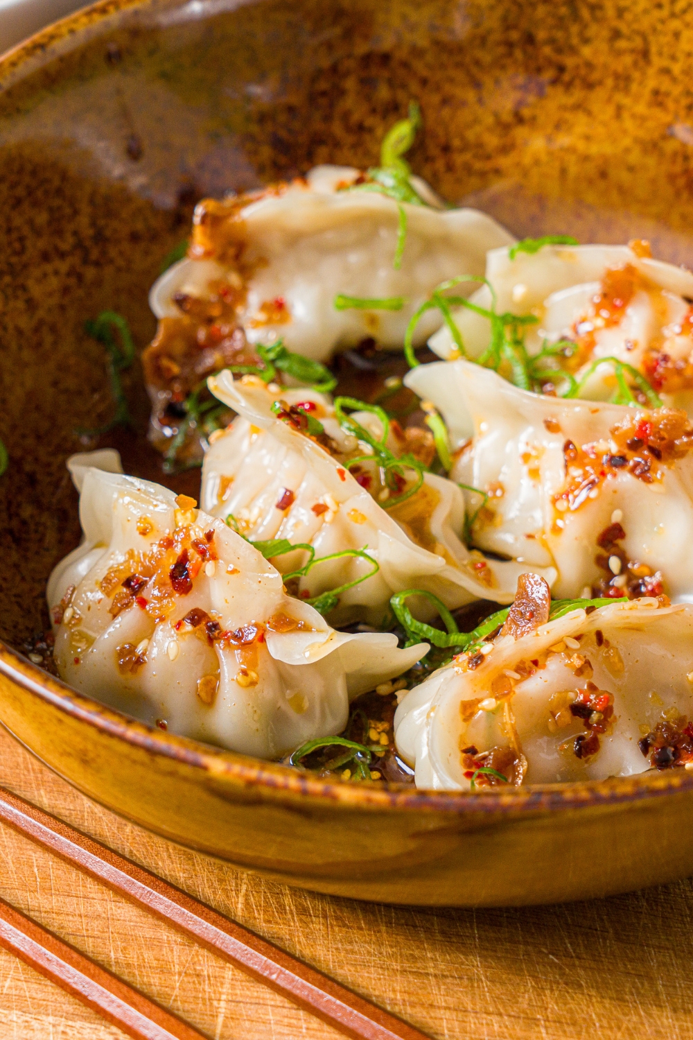 A bowl of steamed pork dumplings in chili oil topped with sliced green onion. The bowl is on a wooden board with a pair of chopsticks.
