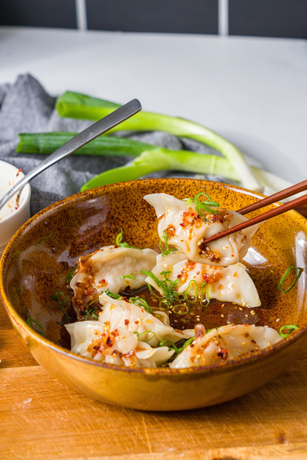 A bowl of steamed pork dumplings in chili oil topped with sliced green onion. A pair of chopsticks is taking one of the dumplings. The bowl is on a wooden board.