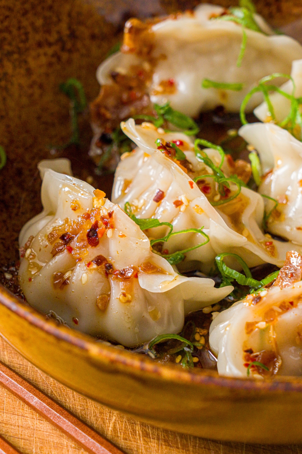 A bowl of steamed pork dumplings in chili oil topped with sliced green onion. The bowl is on a wooden board.
