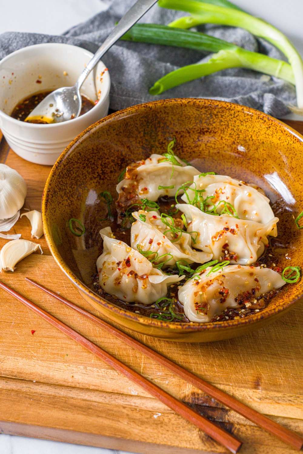 A bowl of steamed pork dumplings in chili oil topped with sliced green onion. The bowl is on a wooden board with a pair of chopsticks and small bowl of chili oil.