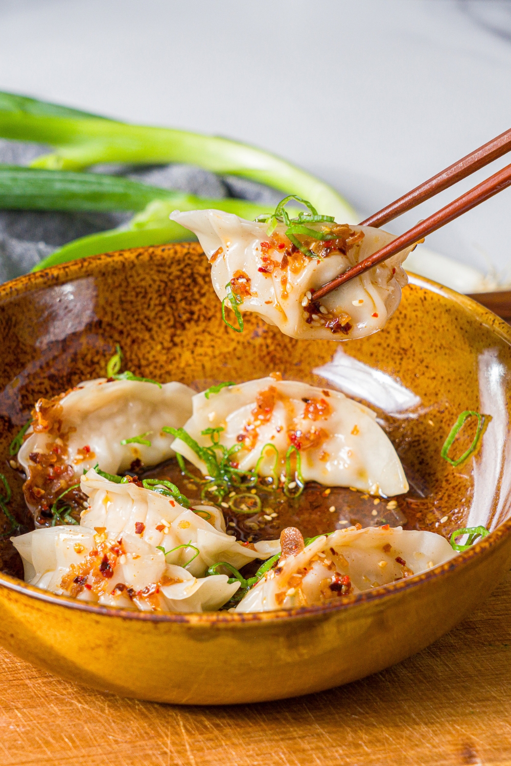 A bowl of steamed pork dumplings in chili oil topped with sliced green onion. A pair of chopsticks is taking one of the dumplings. The bowl is on a wooden board.