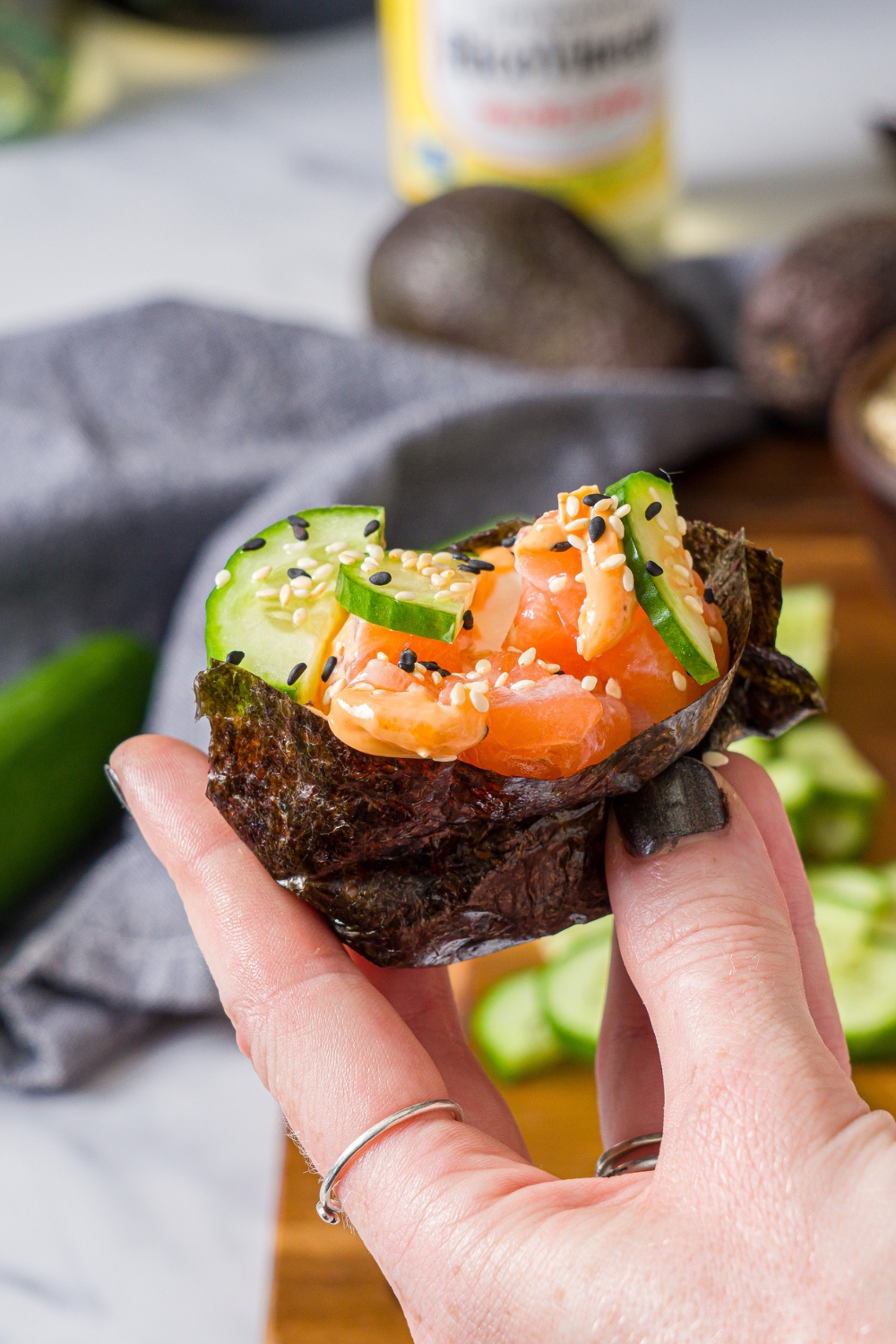 A hand holding a nori sheet stuffed with salmon sushi cake topped iwth sliced cucumber and sesame seeds.