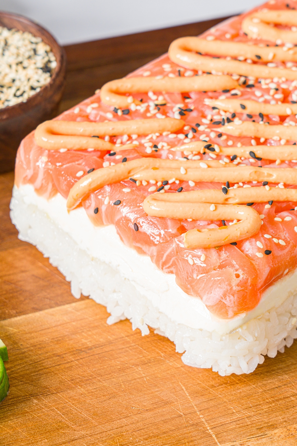 A wooden board with sushi cake with with sushi rice layered with cream cheese and cubed salmon topped with spicy mayo and sesame seeds.