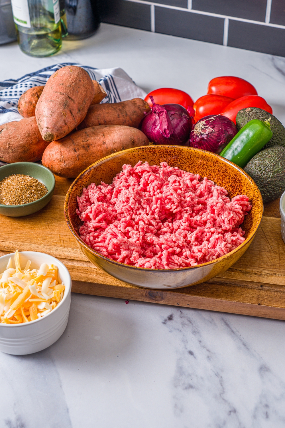 A wooden board with ingredients to make sweet potato nachos including sweet potatoes, ground beef, shredded cheese, avocado, red onion, tomatoes, and seasonings.