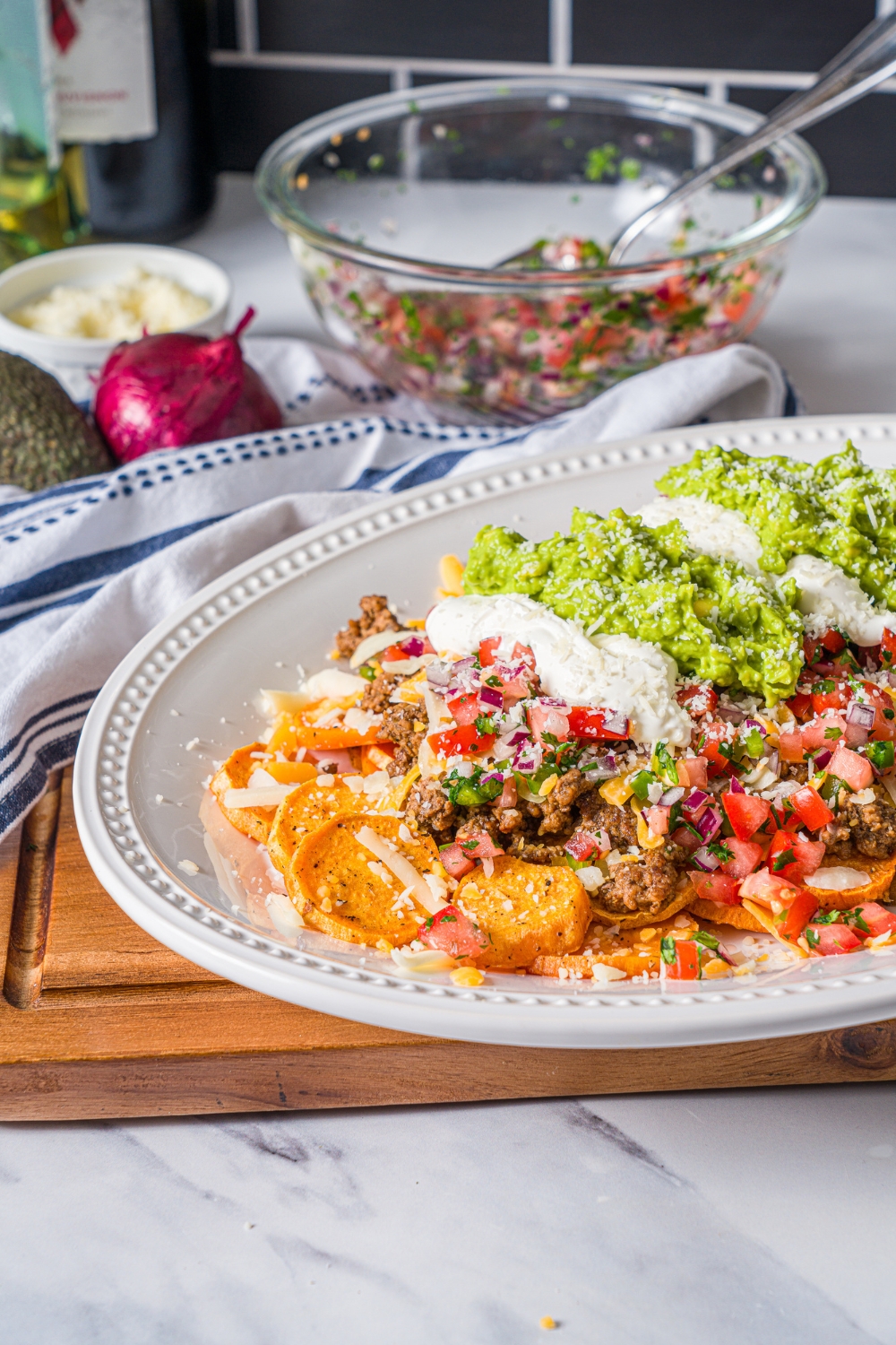 A white platter with sweet potato nachos including sweet potato medallions topped with seasoned ground beef, pico de gallo, cheese, sour cream, and guacamole. The platter is on a wooden board on a marble counter with a blue striped napkin.